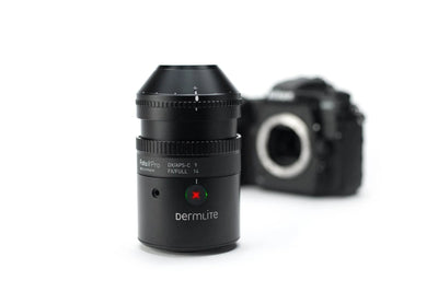 A black Dermlite DVAPS-C 7 full-frame camera lens in sharp focus in the foreground, with a blurred black camera body in the background on a clean white studio backdrop.
